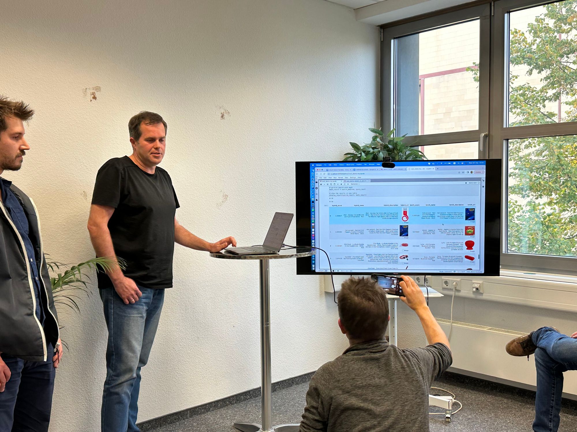 Three individuals engage in a presentation involving a large monitor displaying a webpage, in a room with a whiteboard and a window with greenery