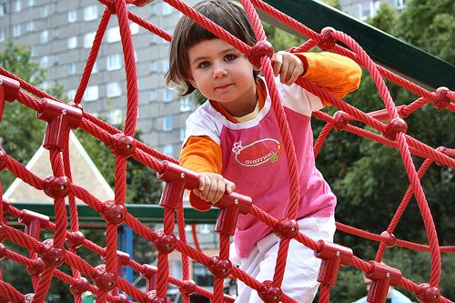 Young girl climbs red rope structure in lively playground, with a building background hinting at urban setting.