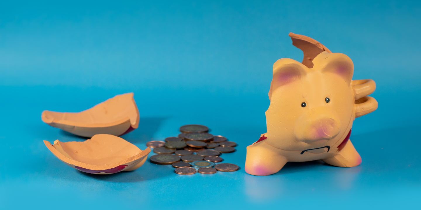 Broken yellow piggy bank on a blue background with scattered coins around it, suggesting financial loss