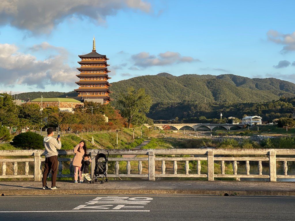 Family by a river with a bridge, admiring a pagoda against a hill under a blue, partly cloudy sky.