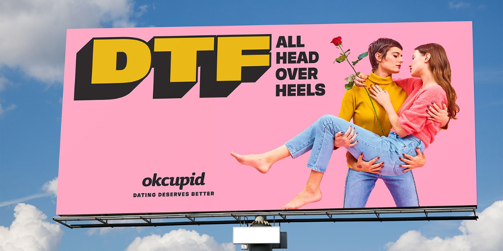 A playful and bold OkCupid billboard advertisement set against a blue sky with white clouds. The ad features a pink background with the tagline "DTF All Head Over Heels" in bright yellow lettering, and depicts two women – one blond and one brunette. The brunette is pictured with a tilted head while the blond woman is holding a red rose. Below them, the phrase "Dating Deserves Better" complements the OkCupid logo