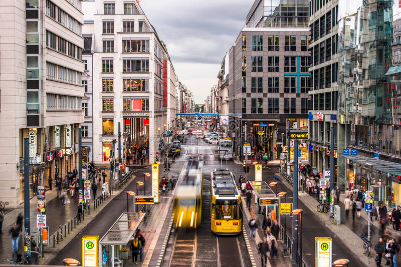 An active urban street in Berlin with tall buildings lining both sides, bustling with pedestrians and street trams. The atmosphere suggests a vibrant city life with people going about their daily routines, reflecting the dynamic energy of a major metropolis