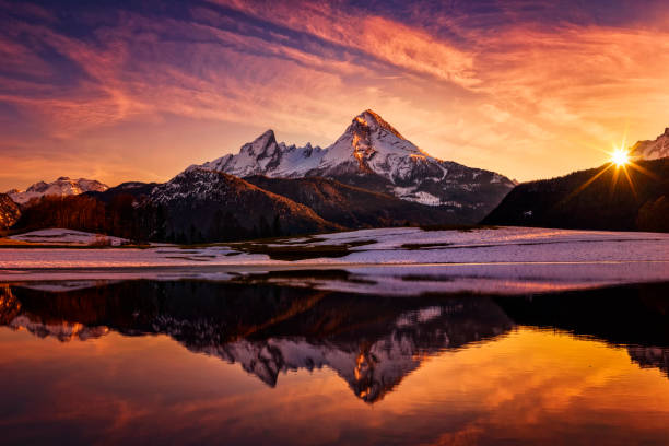 A tranquil winter scene featuring a sunset over a snow-capped mountain range, with the vibrant orange hues of the dusk sky reflecting in the still waters of a lake