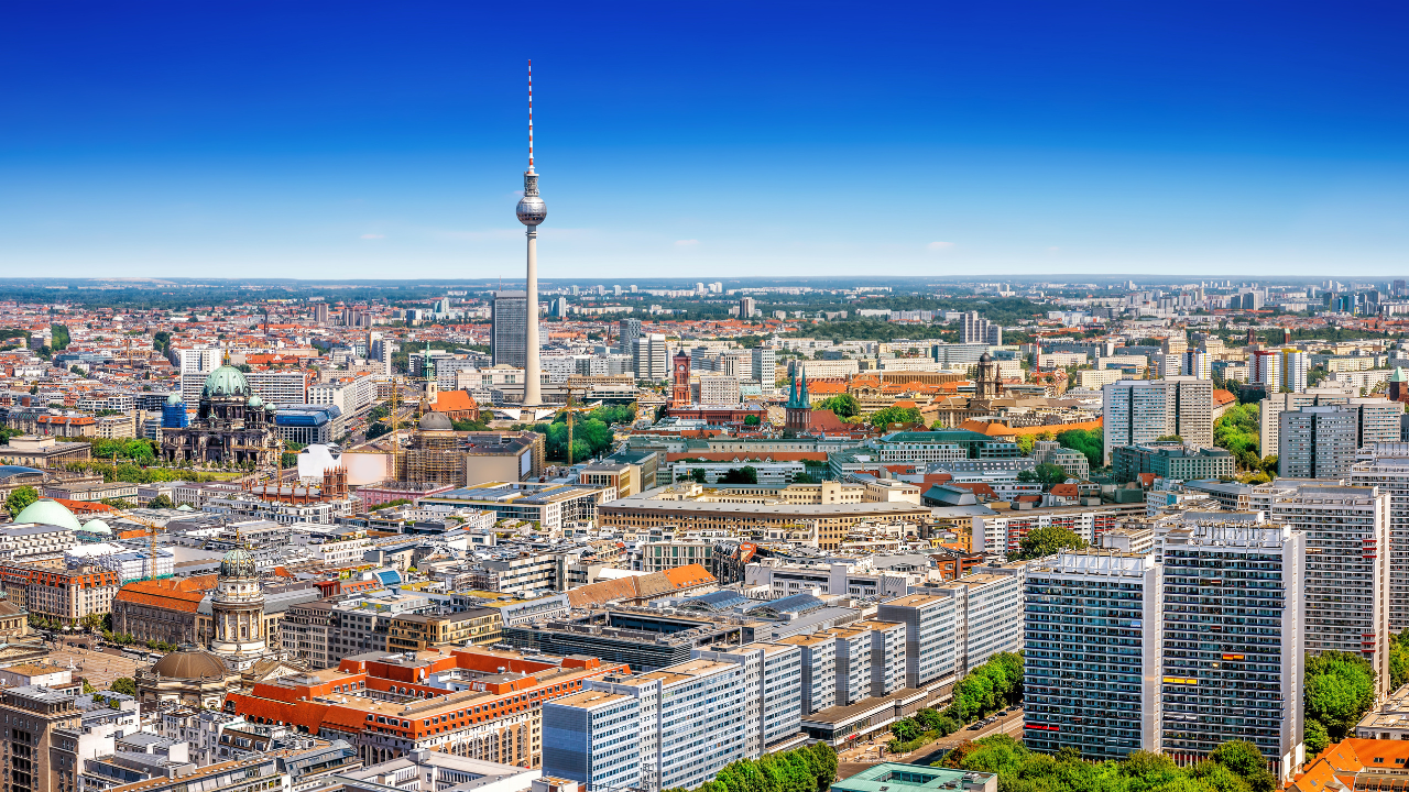 Berlin cityscape featuring the landmark TV Tower, historical red-roofed buildings, and surrounding greenery under a clear blue sky