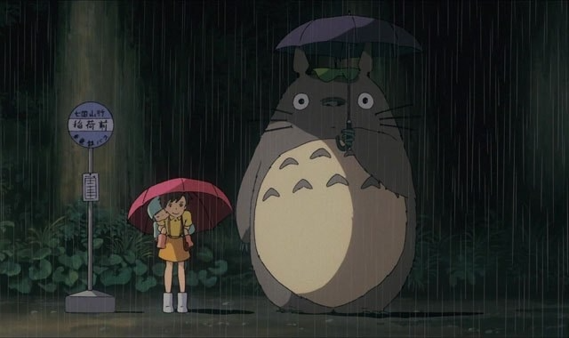 Animated scene featuring Totoro and a young girl in yellow under umbrellas at a bus stop with a dark forest background, from "My Neighbor Totoro.