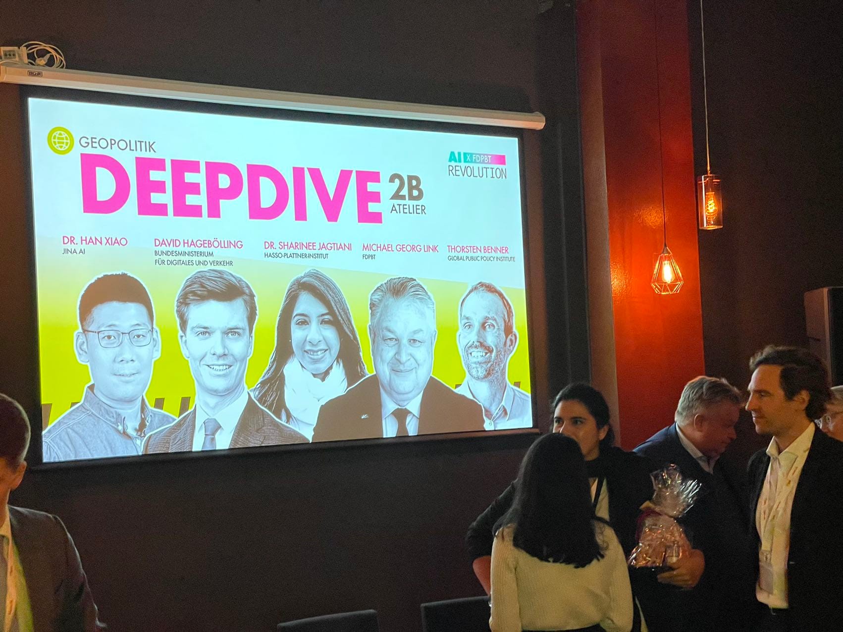 Event attendees in a room with a screen displaying "DeepDive 2B" and multiple speakers, with decor indicating a symposium on geopolitics and digital topics
