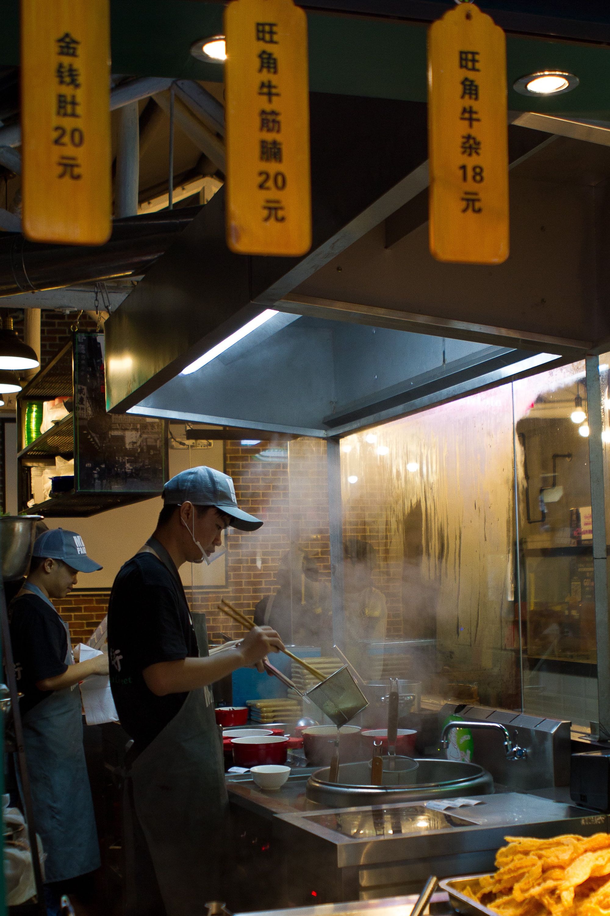 Outdoor Asian food stand with two cooks preparing dishes, utensils on the counter, and visible Chinese dish names with prices