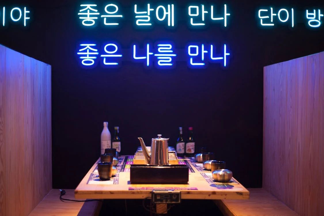 A cozy room with a wooden table, illuminated by a neon sign in Korean and subtle lighting, evoking an intimate, modern vibe