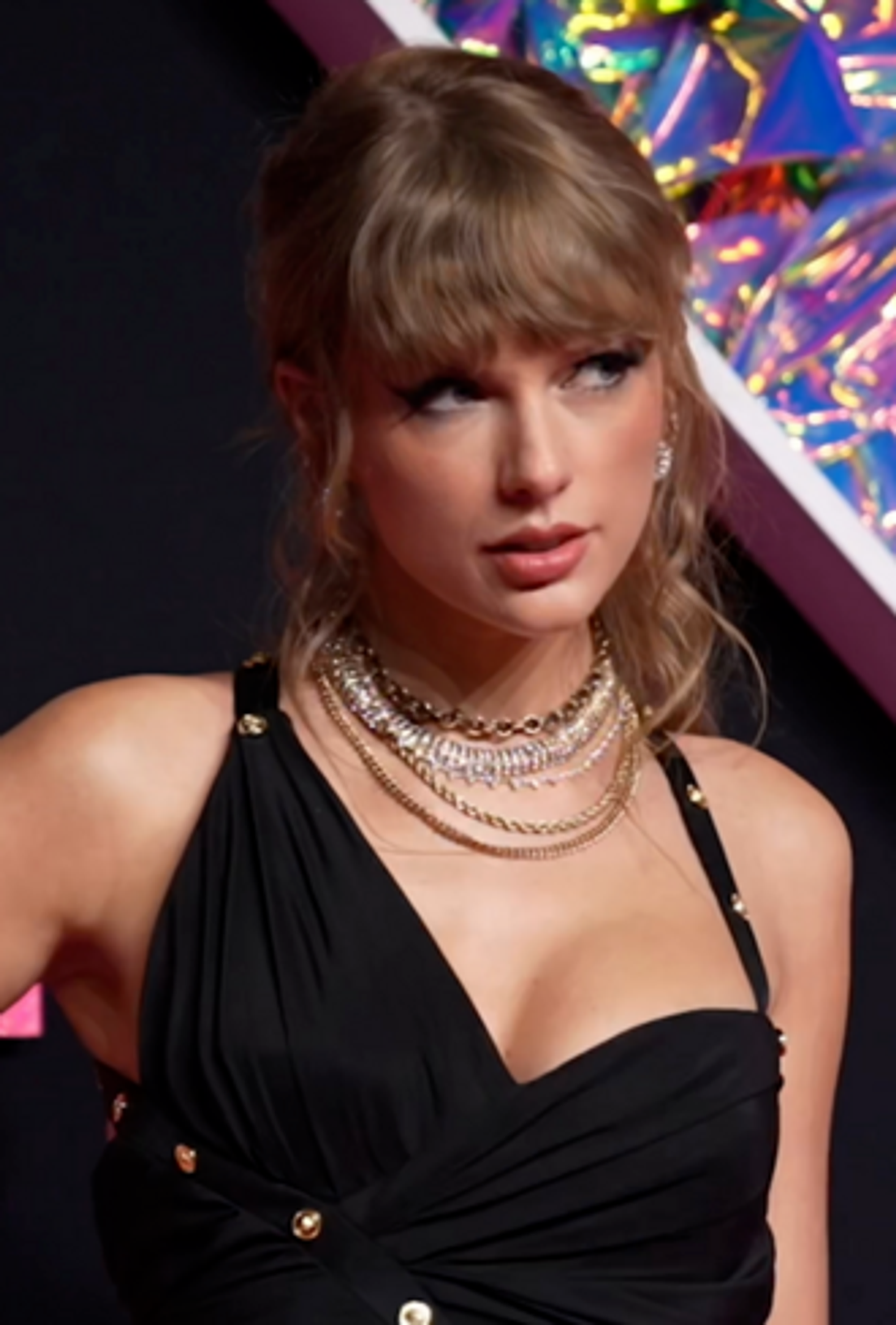 Taylor Swift wearing a black dress with deep V-neckline and gold necklace, posing with a hand on her hip, against a colorful 