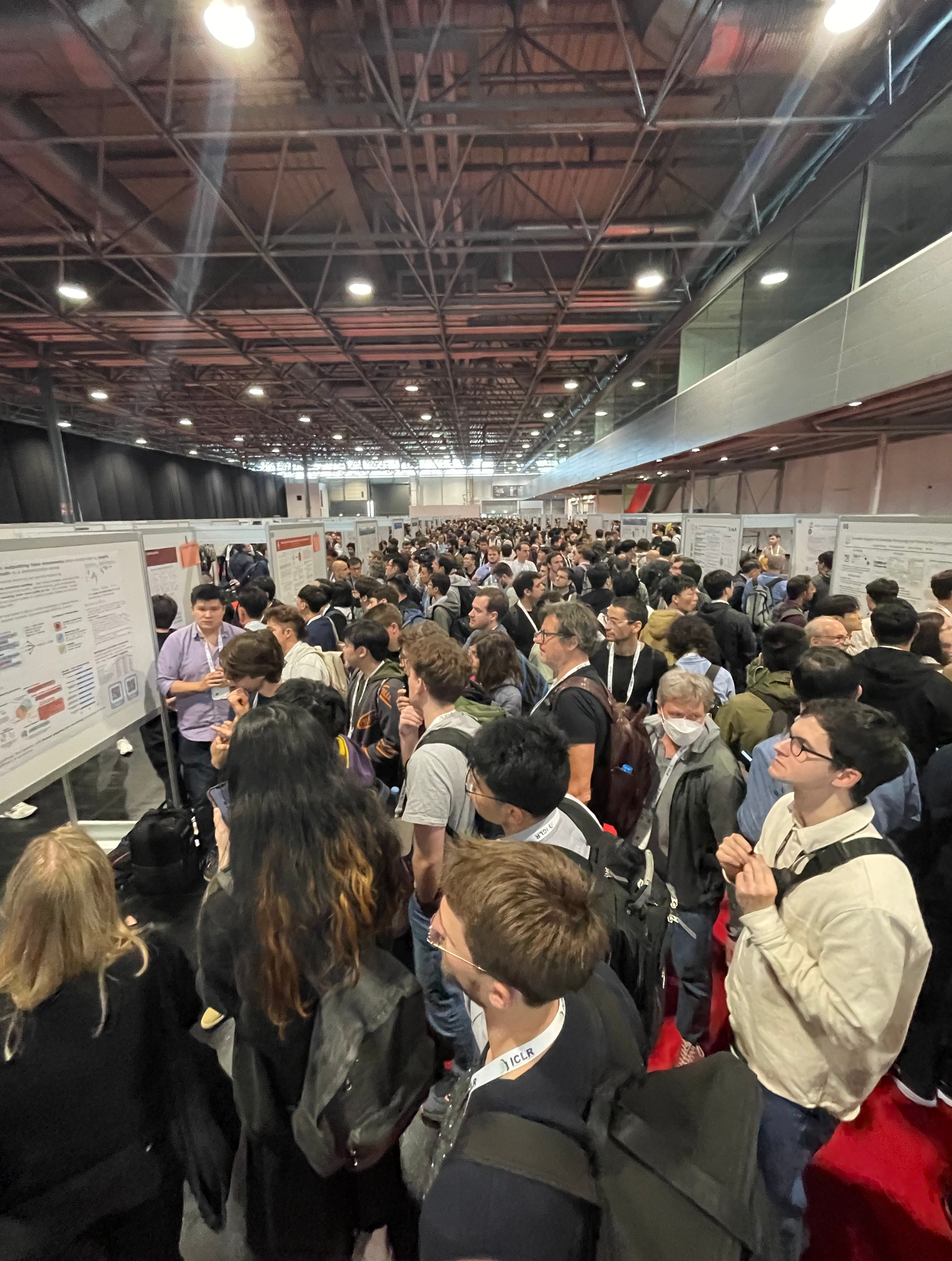 Crowded exhibition hall with people viewing research posters, some wearing lab coats or suits, under a metal truss roof, with