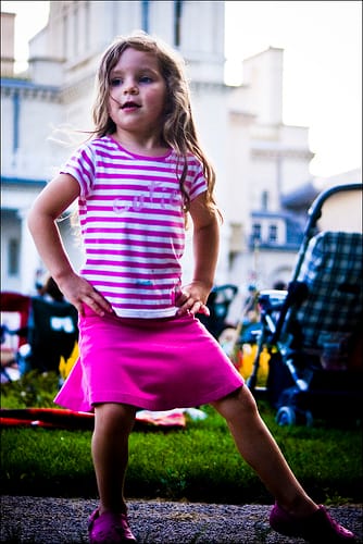 A young girl in a pink skirt playing with a frisbee in an urban outdoor setting with cars and bikes present.